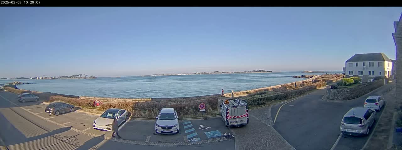A clear, sunny day overlooks a calm body of water with several cars parked in a lot near a building and a small, low wall.
