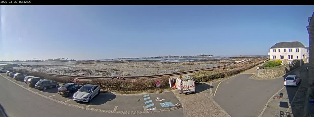 A seaside parking lot with several cars is adjacent to a low stone wall and a stretch of exposed, rocky shore under a clear blue sky.