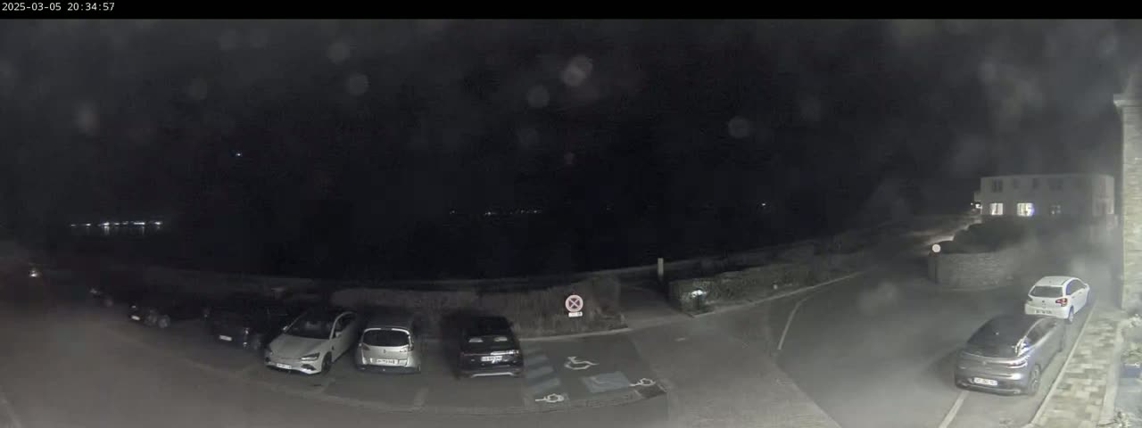 A nighttime view of a parking lot with several cars parked, a building in the background, and a dark, possibly overcast sky.