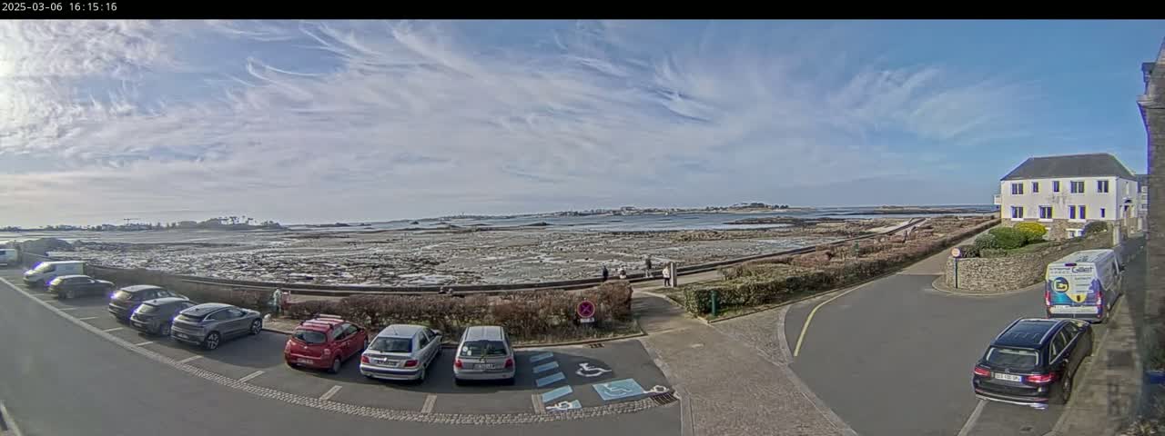A parking lot with several cars is situated near a waterfront area with exposed rocks and a calm sea under a partly cloudy sky.