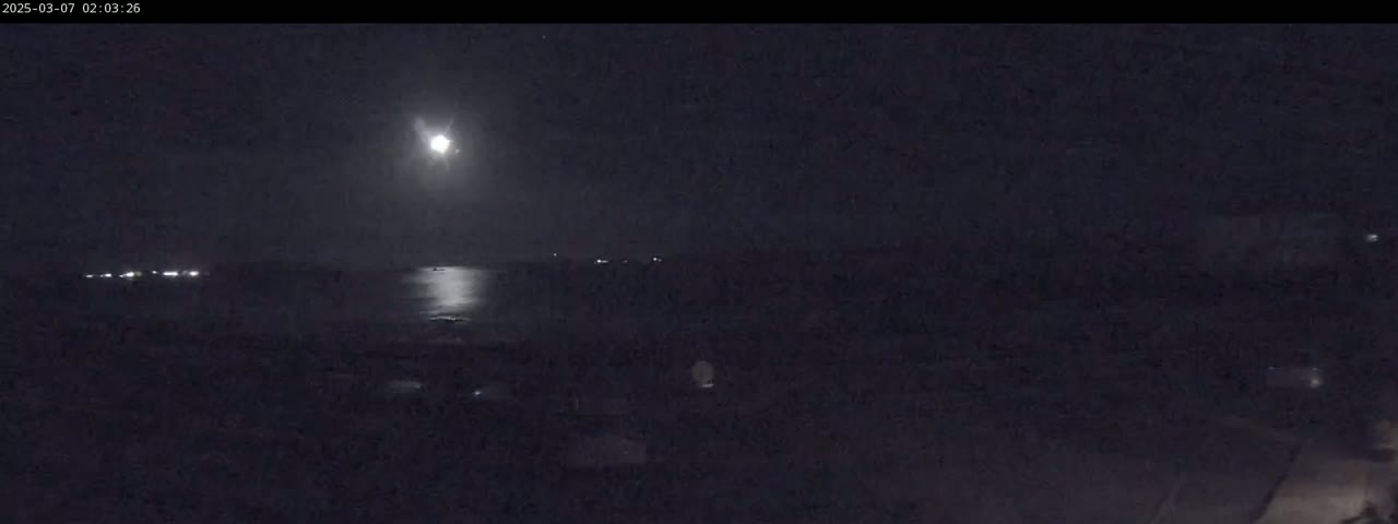 A bright moon illuminates a dark, nighttime scene of water reflecting its light, with distant lights visible across the water and on land.