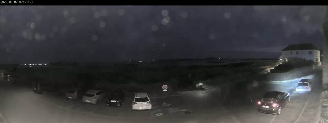 A nighttime, overcast scene shows several cars parked in a lot near a building overlooking a dark body of water.