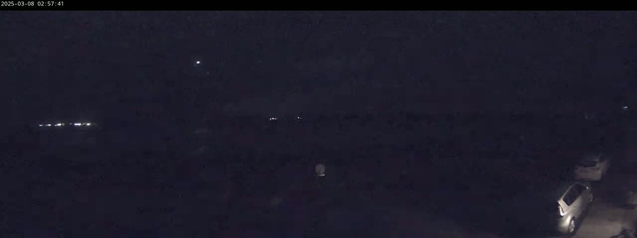 A nighttime, low-light view shows a few parked cars and distant lights.