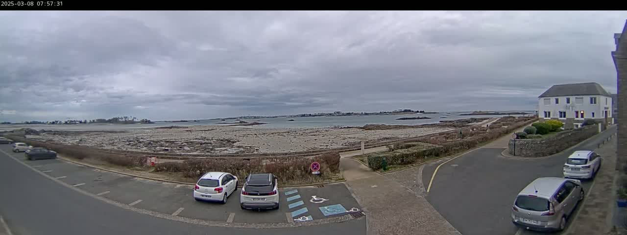 A coastal parking lot with several cars overlooks a rocky beach and calm water under an overcast sky.