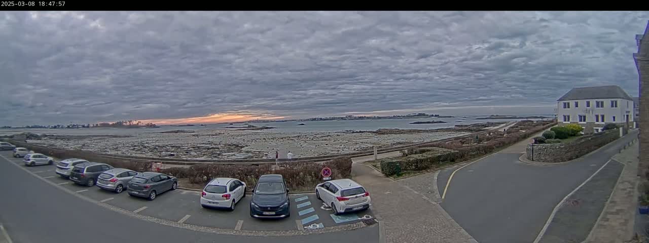 Several cars are parked in a lot overlooking a rocky shoreline and calm body of water under a cloudy sky.