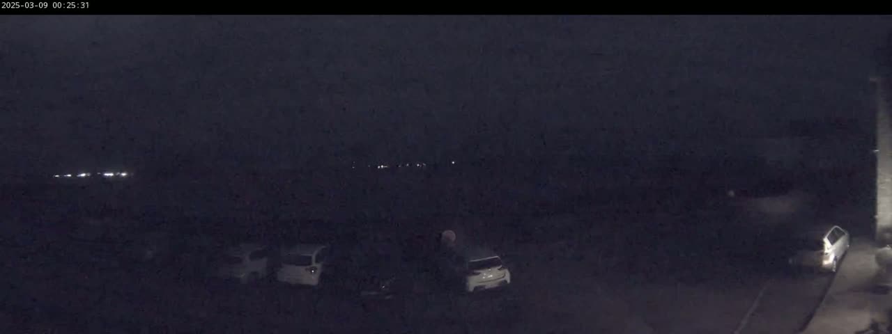 A dark, nighttime scene shows several cars parked in a lot, with faint lights visible in the distance.
