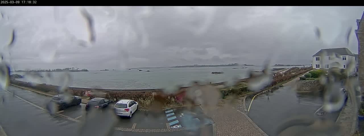 A rainy view of a coastal parking lot with several cars, overlooking a calm sea and distant land under a cloudy sky.