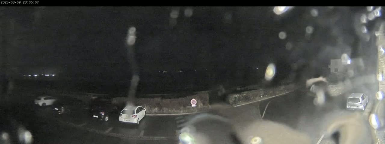 A nighttime outdoor scene shows several cars parked in a lot, viewed through rain-covered glass, with distant lights visible in the background.