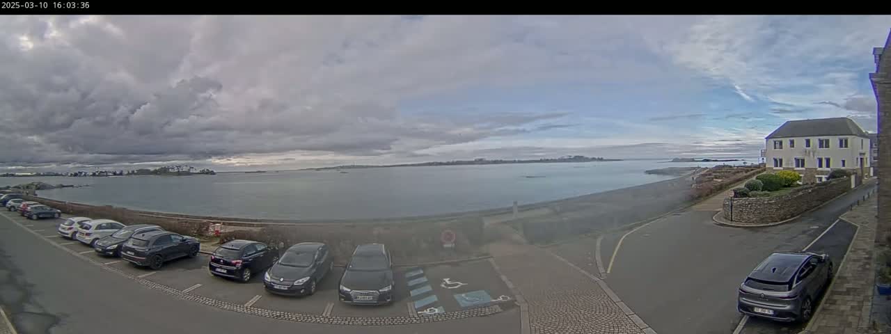 A mostly cloudy day reveals a coastal scene with parked cars near a building overlooking calm water and a distant shoreline.