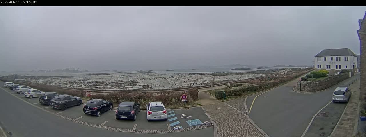 A foggy coastal scene shows a parking lot with several cars next to a road that leads to a building overlooking a rocky shoreline and calm ocean.