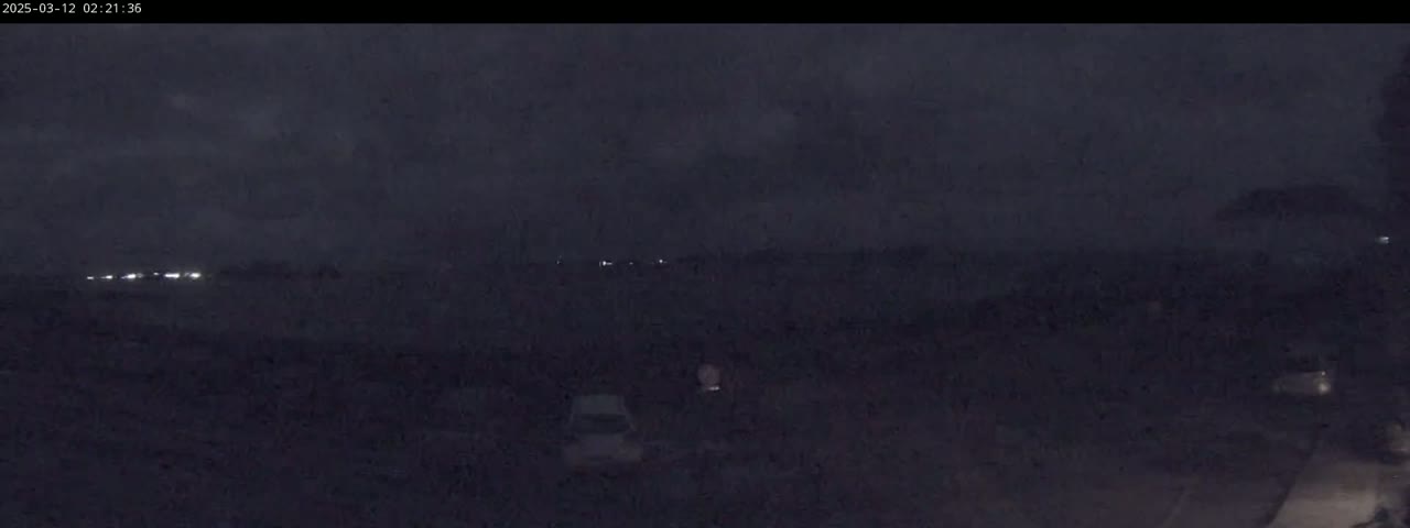 Under a dark, cloudy night sky, a few vehicles are parked in a dimly lit area with distant lights visible on the horizon.