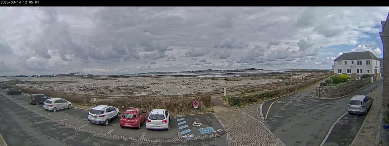 A parking lot with several cars is situated near a coastal area with a rocky shore under a cloudy sky.