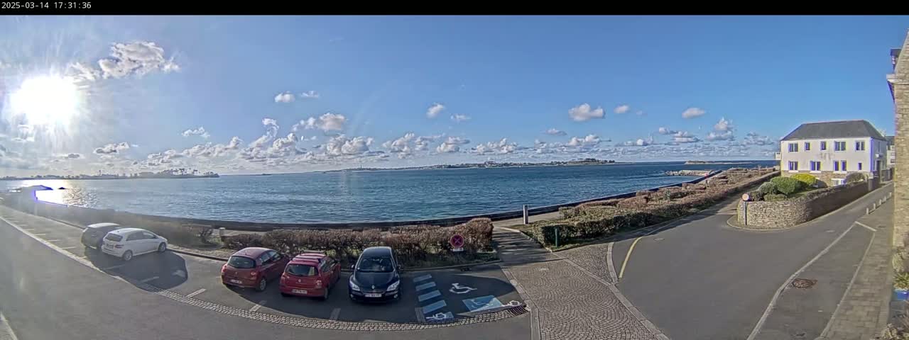A sunny day with partly cloudy skies shows a body of water with a shoreline featuring a parking lot with several cars, landscaping, and a building.