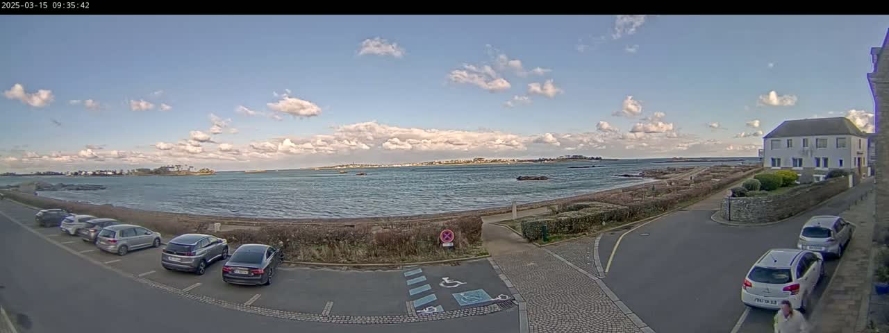 A partly cloudy day reveals a tranquil coastal scene with parked cars near a waterfront road and a building overlooking a calm body of water.