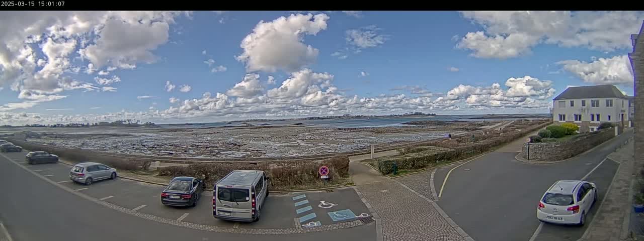 A partly cloudy day shows a coastal scene with a parking lot containing several cars and a van, overlooking a wide expanse of tidal flats leading to a body of water and a distant shoreline.