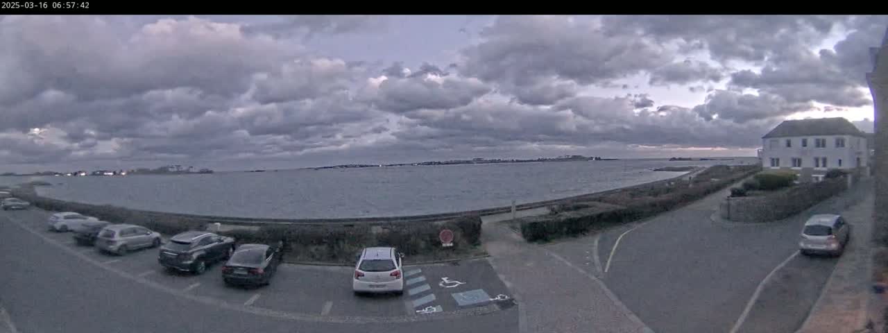 Several cars are parked in a lot overlooking a calm body of water under a cloudy sky.