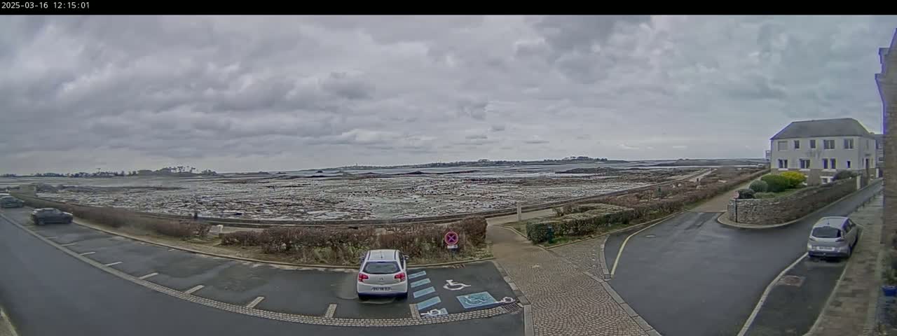A parking lot with a few cars is situated near a coastal area with overcast skies and low-lying land that appears to be exposed at low tide.