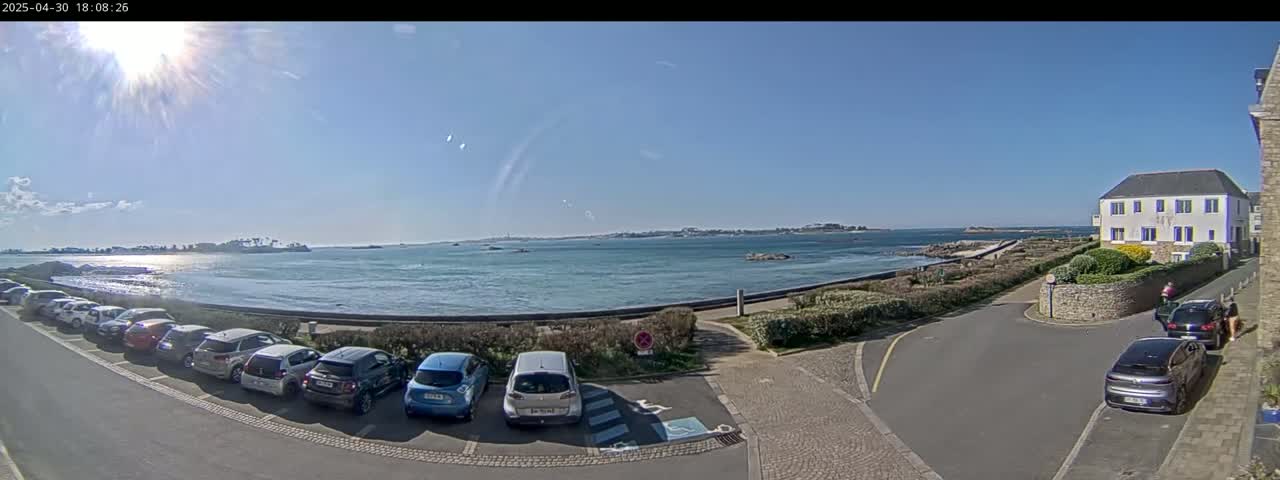 A sunny day shows a parking lot full of cars next to a calm body of water with a distant coastline under a mostly clear sky.