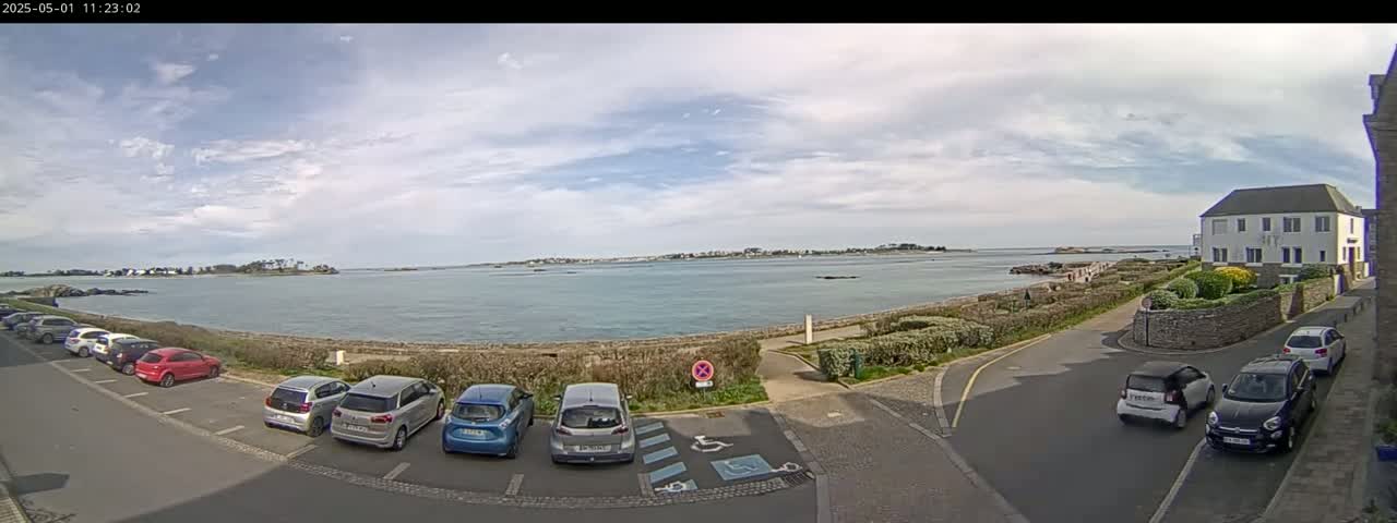 A mostly sunny day shows a calm body of water with a distant shoreline, several cars parked in a lot, and a residential building.