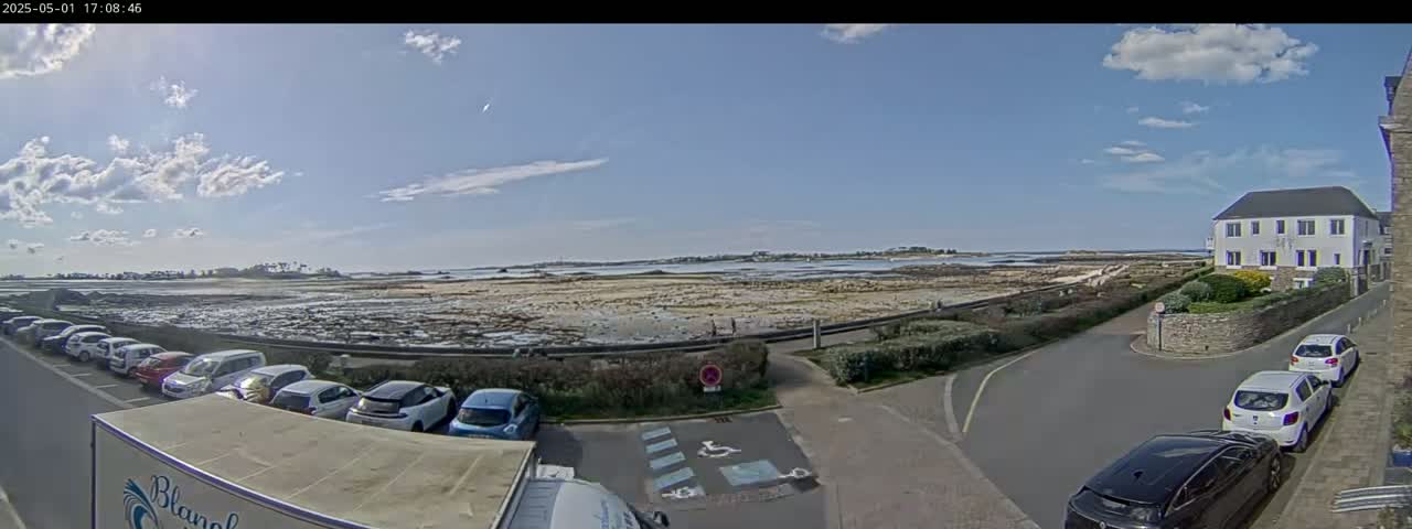 A partly cloudy day reveals a coastal scene with a parking lot full of cars near a road leading to a calm body of water, extending to a distant shoreline.