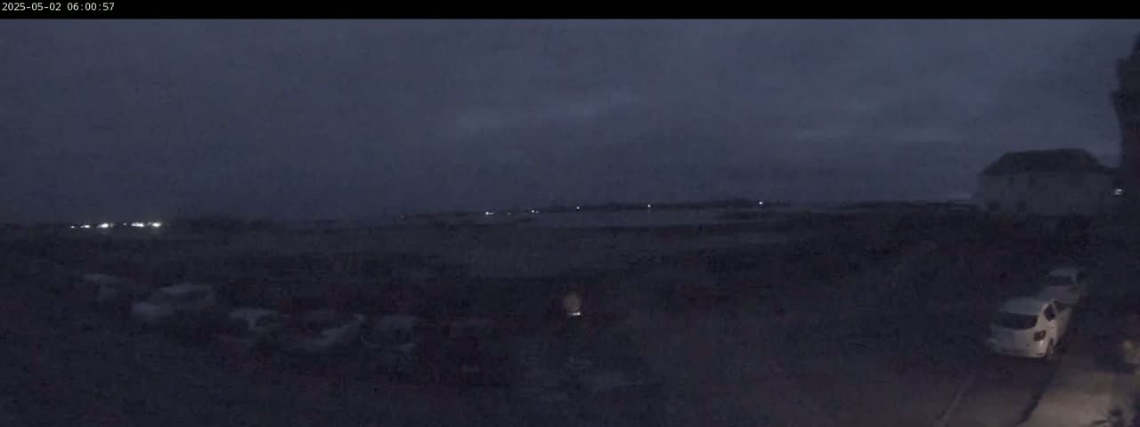 Under a dark, overcast sky, several vehicles are parked in a lot near a building, with a few more headlights visible in the distance.