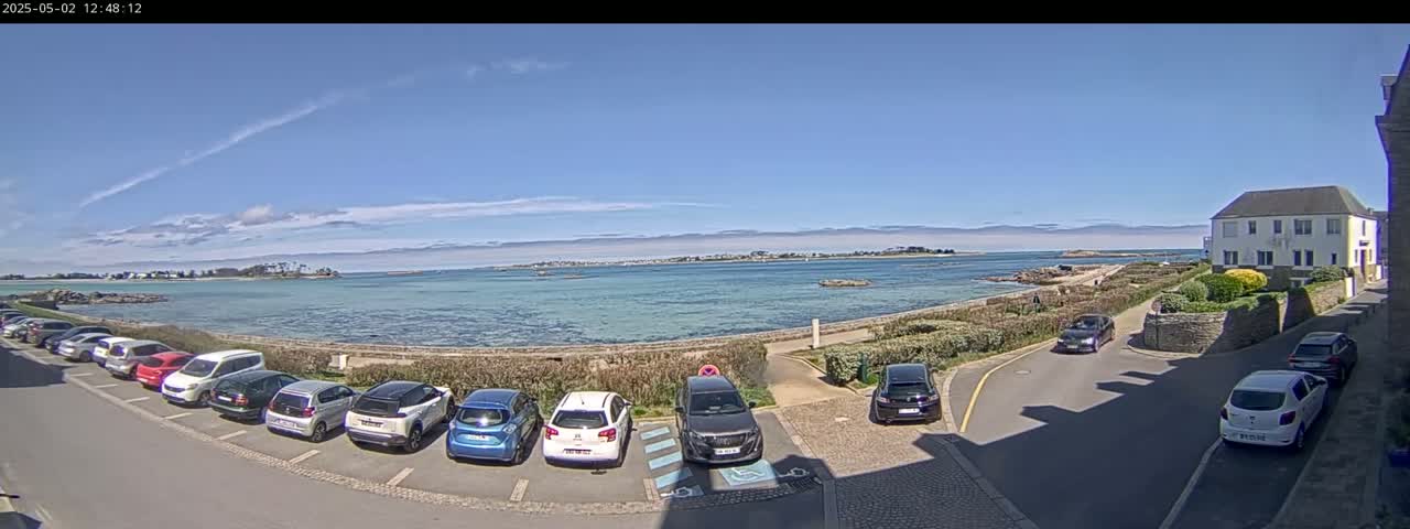 A sunny day reveals a coastal scene with parked cars lining a street next to a calm body of water, a distant shoreline, and a house with a white exterior.