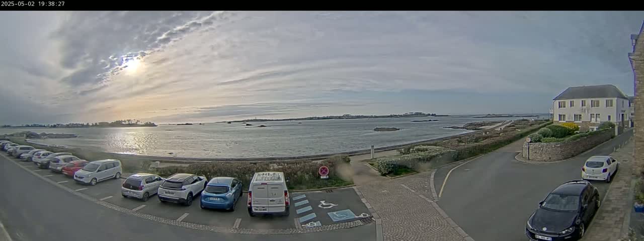 A partly sunny day shows a calm body of water with a distant shoreline, a parking lot with several cars, and a building on the right.