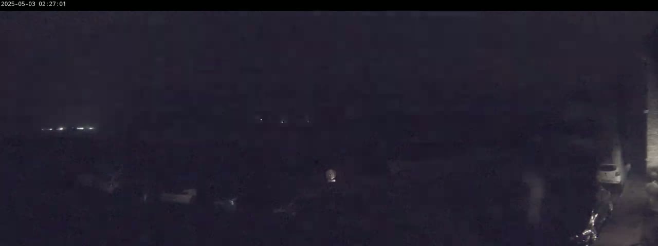 A dark, nighttime scene shows indistinct shapes and faint lights in the distance under a mostly clear sky.
