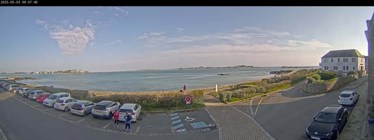 A partly cloudy day shows a coastal parking lot with several cars, a view of calm water and a distant shoreline, and a white building near a stone wall.