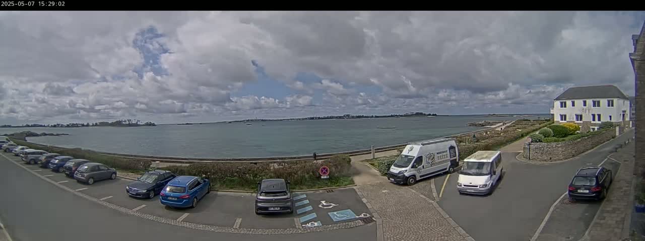 A partly cloudy day shows a seaside parking lot with several cars, vans, and a building overlooking calm water and a distant shoreline.