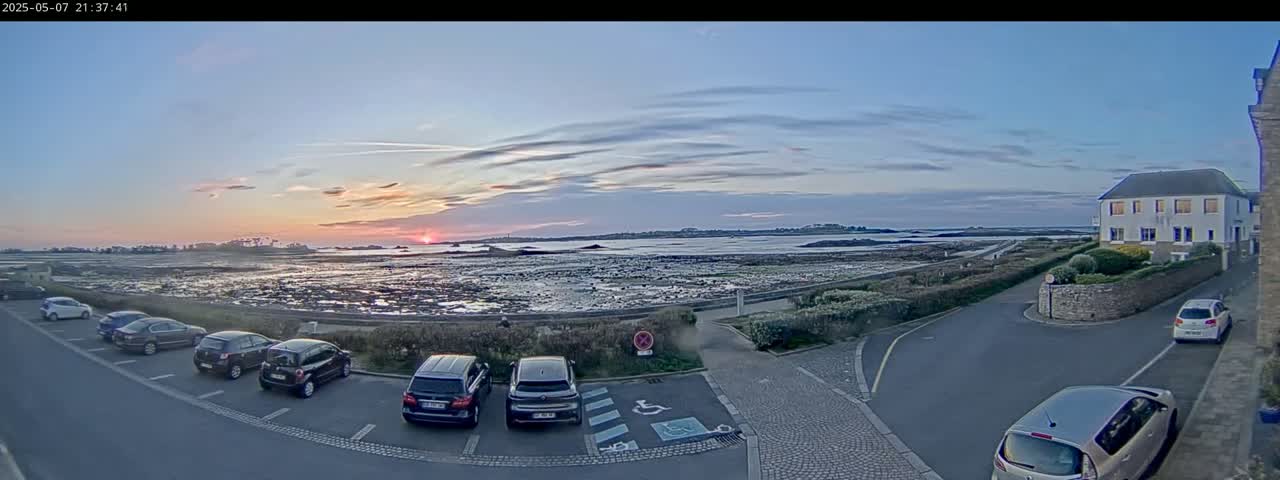 Several cars are parked in a lot overlooking a calm body of water at sunset under a partly cloudy sky.