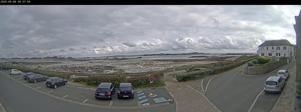 A parking lot with several cars is next to a road overlooking a wide expanse of tidal flats under a cloudy sky.