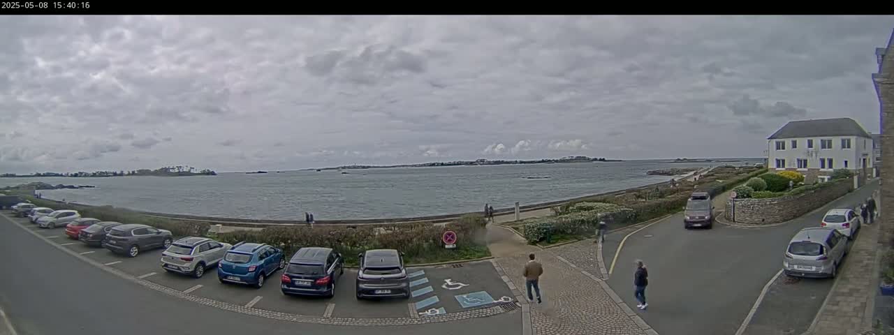 A parking lot next to a calm body of water under a cloudy sky, with several cars parked and a few people walking nearby.