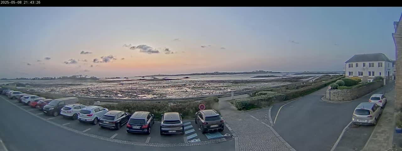 A parking lot with several cars is situated near a body of water at sunset, under a mostly clear sky.