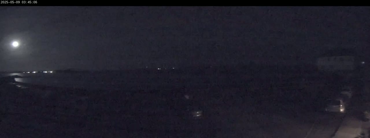 A bright moon illuminates a dark, nighttime scene of water with distant lights and land with some vehicles visible.