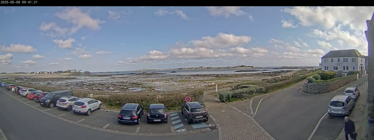 A partly cloudy day reveals a coastal scene with a parking lot full of cars near a building overlooking a rocky shoreline and calm body of water.