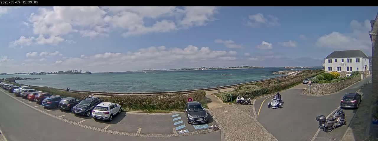 A partly cloudy day shows a coastal parking lot with several cars, a few motorcycles, and a three-wheeled vehicle near a white building overlooking a calm body of water and a distant landmass.