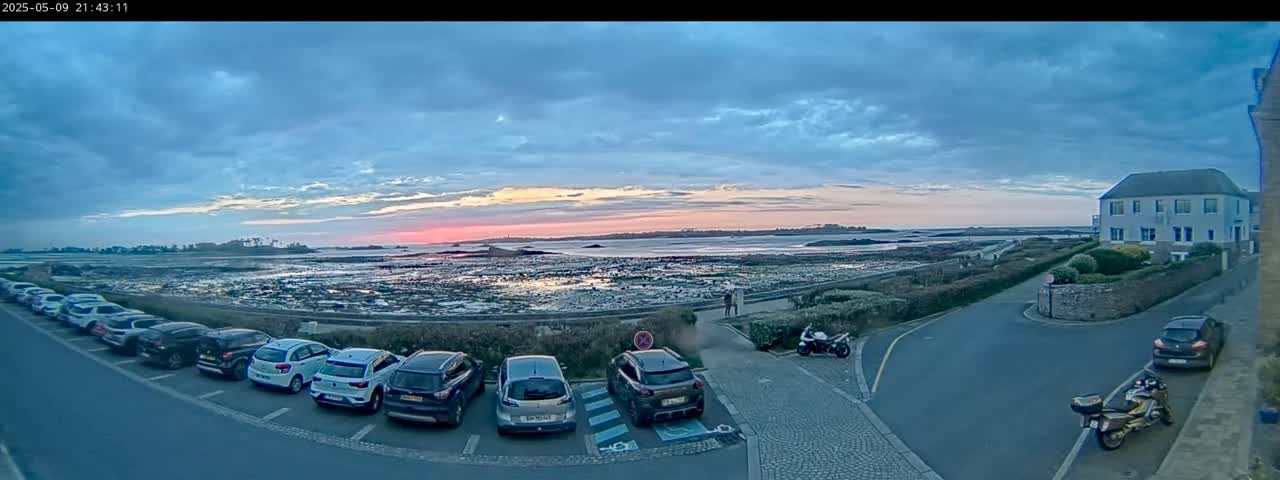 A parking lot with several cars is situated next to a body of water under a twilight sky with a pink and orange sunset.