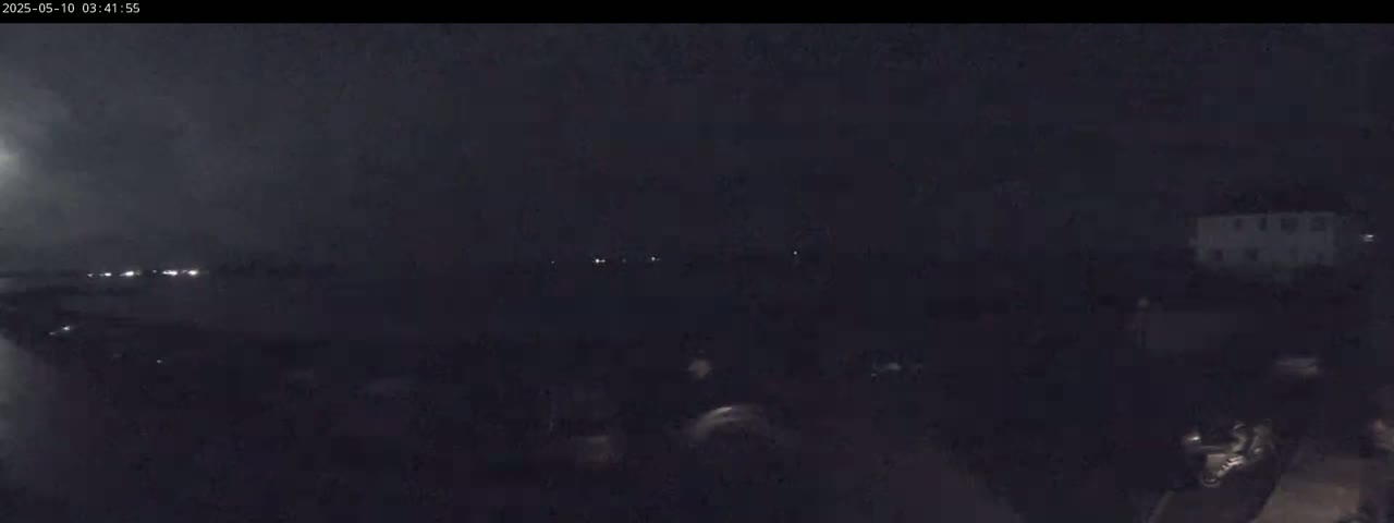 A dark, nighttime scene shows a few distant lights and indistinct shapes in the foreground, under a cloudy sky.