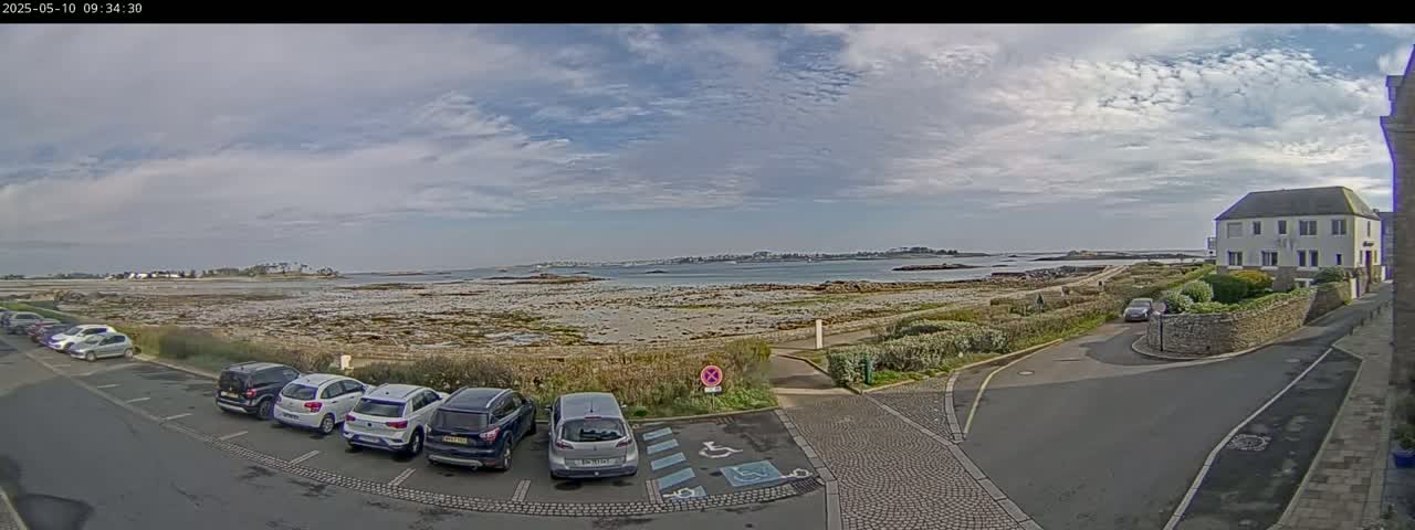 A parking lot with several cars is next to a road leading to a calm body of water under a partly cloudy sky.