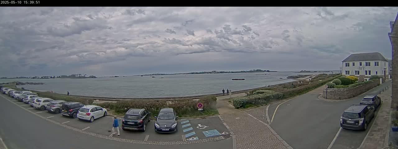 A parking lot with several cars overlooks a calm body of water under a cloudy sky, with a small town visible in the background.