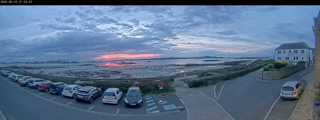 A row of cars is parked in a lot overlooking a calm body of water under a partly cloudy sky with a pinkish sunset.