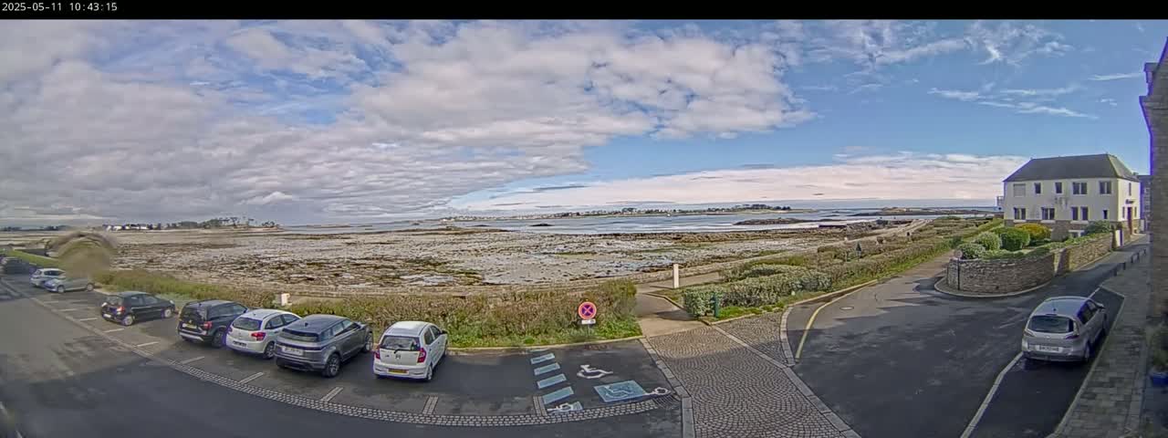 A partly cloudy day reveals a coastal scene with parked cars near a building, overlooking a rocky shoreline and calm body of water.