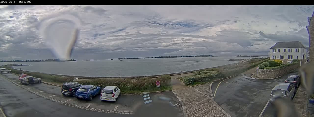 A coastal scene shows overcast skies, a calm body of water, several cars parked in a lot near a building, and wet roads.