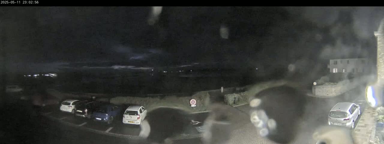 A nighttime view of a parking lot with several cars parked, under a dark, cloudy sky with some rain or moisture on the camera lens.