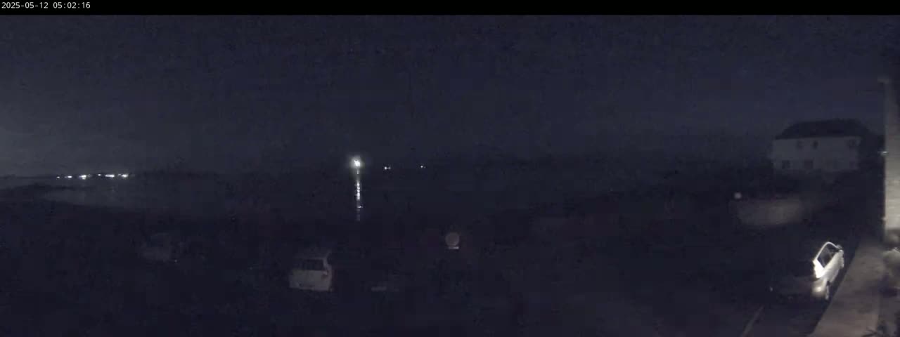 A nighttime, dark scene shows a body of water in the distance with faint lights, a few parked cars, and a building in the foreground under a mostly clear, dark sky.