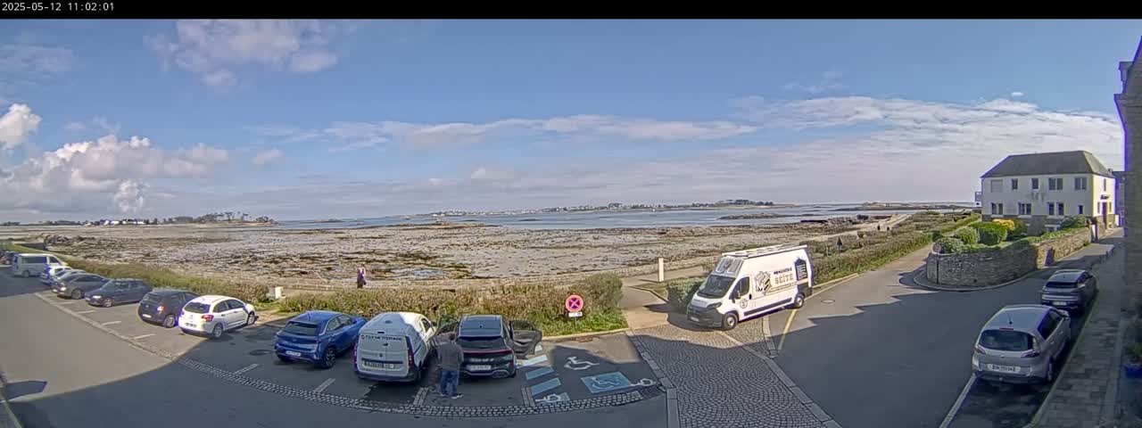 A seaside parking lot with several cars parked, next to a calm body of water under a partly cloudy sky.
