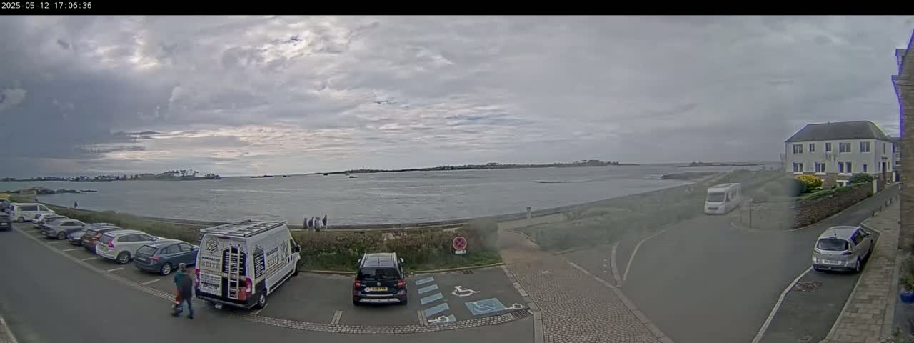 A mostly cloudy day shows a calm body of water with a shoreline featuring parked cars, a van, and a building, with a few people standing near the water.
