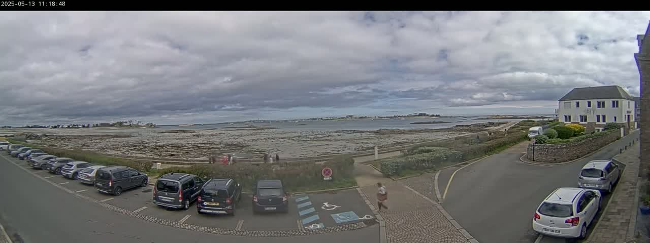A seaside parking lot with several cars is visible under a cloudy sky, overlooking a rocky shoreline and calm water.