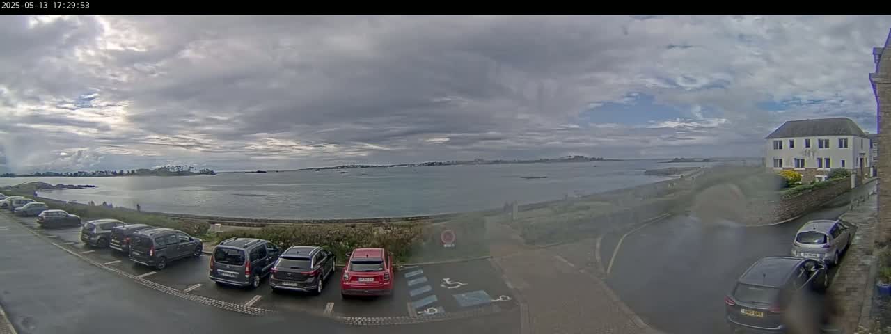 A parking lot with several cars is situated near a calm body of water under a mostly cloudy sky.